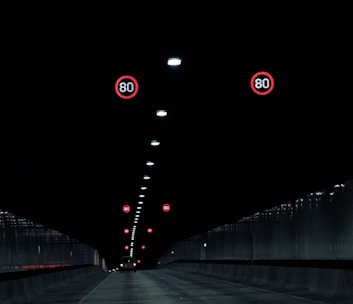 A dark tunnel interior with illuminated speed limit signs indicating 80 km/h. The tunnel is lined with ceiling lights running down the center, and a single vehicle is visible in the distance moving away.