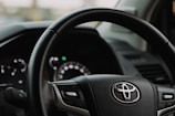 Close-up view of a Toyota steering wheel inside a vehicle, showing the brand logo prominently in the center. The dashboard and part of the driver's side door are visible in the background.