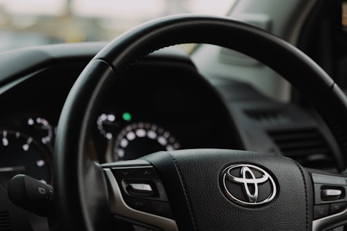Close-up view of a Toyota steering wheel inside a vehicle, showing the brand logo prominently in the center. The dashboard and part of the driver's side door are visible in the background.