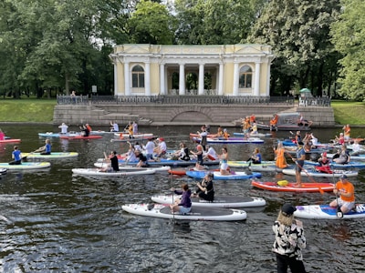 A large group of people are paddleboarding on a river in front of a classical pavilion surrounded by lush green trees. The diverse crowd includes individuals of various ages, all engaged in the activity with some wearing vibrant clothing and life vests. A woman in the foreground is taking photos or videos.