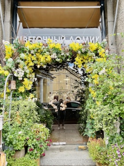 An inviting entrance adorned with a lush, vibrant floral arrangement that includes yellow and white flowers. The space is surrounded by various green plants and potted flowers, creating a welcoming and natural atmosphere. A reflection on a glass door shows a person standing outside, contributing to the bustling urban environment.