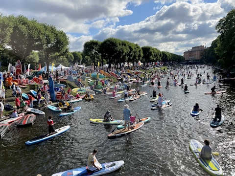 A bustling scene on a river with numerous people paddleboarding. The river is flanked by lush, green trees and a large crowd gathered on the bank. Paddleboards of various colors and sizes fill the water, and the atmosphere is lively with activity. A building can be seen in the distance under a sky dotted with clouds.