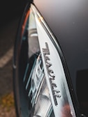 Close-up view of a car headlight featuring the Maserati logo, with sleek and glossy metallic surfaces reflecting light.