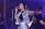 A singer passionately performs on stage, holding a microphone. She is wearing a shiny, metallic outfit and large hoop earrings. Her hair is in a ponytail, and she extends one arm wide. Beside her, a band member wearing glasses and a t-shirt with text is slightly turned away, playing an instrument. The background features stage lighting with a purple hue.