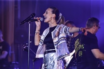 A singer passionately performs on stage, holding a microphone. She is wearing a shiny, metallic outfit and large hoop earrings. Her hair is in a ponytail, and she extends one arm wide. Beside her, a band member wearing glasses and a t-shirt with text is slightly turned away, playing an instrument. The background features stage lighting with a purple hue.