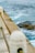 A coastal scene featuring a concrete seawall with metal railings in the foreground. The ocean beyond is a vibrant blue-green, with visible waves and distant kelp. A rocky outcrop covered with seaweed is partially submerged in the water.