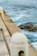 A coastal scene featuring a concrete seawall with metal railings in the foreground. The ocean beyond is a vibrant blue-green, with visible waves and distant kelp. A rocky outcrop covered with seaweed is partially submerged in the water.