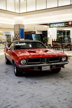 A classic red muscle car with a black roof is parked indoors on a carpeted area. The car displays a license plate with Arabic and English text. In the background, there are decorative elements and several seating arrangements, with signage indicating a café or restaurant named 'Künefeci'. The setting suggests a commercial space or shopping mall with modern and stylish interior design.