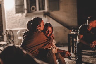 Two people are sitting close together, with one person embracing the other, conveying a sense of warmth and affection. The setting appears to be outdoors at night, illuminated by soft lighting. Another person is seated nearby, suggesting a social gathering.