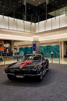 A classic black car with a red detail is displayed indoors on a gray carpeted area. The area is well-lit with modern interior lighting, featuring decorative walls with teal wave patterns. Several people stand nearby, some observing the car.