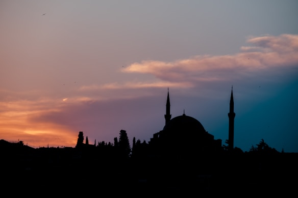 The silhouette of a mosque with two minarets set against a dramatic sky during sunset, with orange and pink hues blending into a darker blue. Trees and building outlines are visible in the foreground.