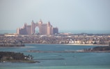 A grand, palatial hotel with pinkish hues stands majestically along the coastline. Surrounding the hotel are numerous luxury villas and buildings, set against the backdrop of a serene blue sea. The sky is hazy, suggesting a warm day by the sea.