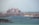 A grand, palatial hotel with pinkish hues stands majestically along the coastline. Surrounding the hotel are numerous luxury villas and buildings, set against the backdrop of a serene blue sea. The sky is hazy, suggesting a warm day by the sea.