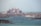 A grand, palatial hotel with pinkish hues stands majestically along the coastline. Surrounding the hotel are numerous luxury villas and buildings, set against the backdrop of a serene blue sea. The sky is hazy, suggesting a warm day by the sea.