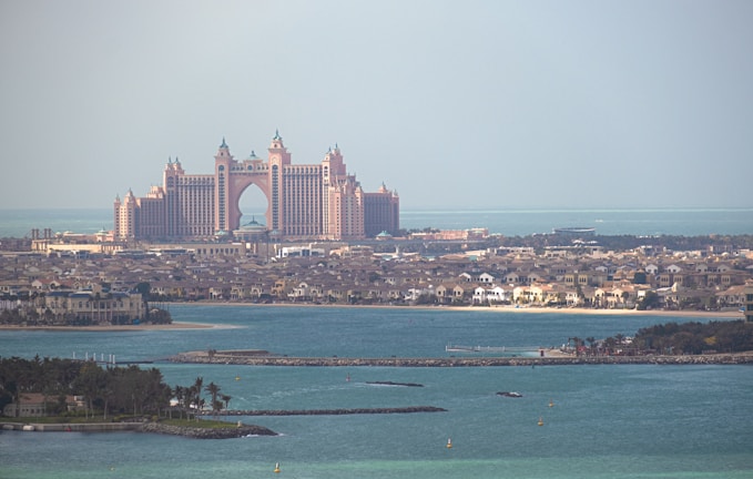 A grand, palatial hotel with pinkish hues stands majestically along the coastline. Surrounding the hotel are numerous luxury villas and buildings, set against the backdrop of a serene blue sea. The sky is hazy, suggesting a warm day by the sea.