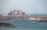 A grand, palatial hotel with pinkish hues stands majestically along the coastline. Surrounding the hotel are numerous luxury villas and buildings, set against the backdrop of a serene blue sea. The sky is hazy, suggesting a warm day by the sea.