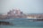 A grand, palatial hotel with pinkish hues stands majestically along the coastline. Surrounding the hotel are numerous luxury villas and buildings, set against the backdrop of a serene blue sea. The sky is hazy, suggesting a warm day by the sea.