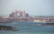 A grand, palatial hotel with pinkish hues stands majestically along the coastline. Surrounding the hotel are numerous luxury villas and buildings, set against the backdrop of a serene blue sea. The sky is hazy, suggesting a warm day by the sea.
