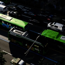 A bustling urban street scene with several vehicles, including bright green buses and a white van, traveling on a multi-lane road. Shadows from nearby buildings create interesting patterns on the road, and the scene appears to take place during daylight hours.