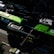 A bustling urban street scene with several vehicles, including bright green buses and a white van, traveling on a multi-lane road. Shadows from nearby buildings create interesting patterns on the road, and the scene appears to take place during daylight hours.
