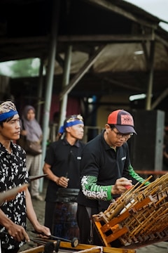 Several individuals in traditional attire are playing musical instruments, including an angklung. They appear to be participating in a cultural performance or rehearsal, with their focus directed at the instruments. The surrounding environment suggests an outdoor setting, perhaps a market or a communal area.