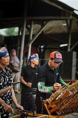 Several individuals in traditional attire are playing musical instruments, including an angklung. They appear to be participating in a cultural performance or rehearsal, with their focus directed at the instruments. The surrounding environment suggests an outdoor setting, perhaps a market or a communal area.