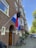 A Dutch flag with a black backpack hanging from the pole is displayed outside a brick apartment building. Several bicycles are parked near the entrance, and the street is lined with trees and parked cars.