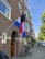 A Dutch flag with a black backpack hanging from the pole is displayed outside a brick apartment building. Several bicycles are parked near the entrance, and the street is lined with trees and parked cars.