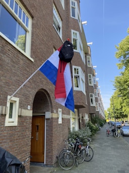 A Dutch flag with a black backpack hanging from the pole is displayed outside a brick apartment building. Several bicycles are parked near the entrance, and the street is lined with trees and parked cars.