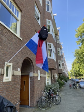 A Dutch flag with a black backpack hanging from the pole is displayed outside a brick apartment building. Several bicycles are parked near the entrance, and the street is lined with trees and parked cars.