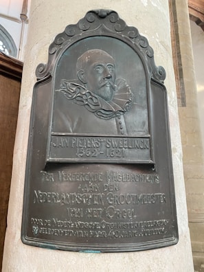 A decorative plaque mounted on a wall depicts a relief portrait of an older man with a ruffled collar. The plaque includes inscriptions in Dutch, commemorating Jan Pieterszoon Sweelinck, a historical figure from 1562-1621.