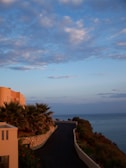 A coastal scene with a modern villa to the left, featuring palm trees and a winding road leading towards the sea. The ocean is visible on the right, and the sky is filled with scattered clouds reflecting a warm, golden hue from the setting sun.