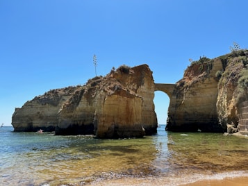 A picturesque coastal scene featuring rugged cliffs with a natural archway. The cliffs are covered with sparse vegetation, and the clear blue sea gently laps against the sandy shore. In the distance, a kayak is visible on the water under a bright blue sky.