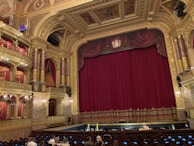 An ornate theatre interior featuring a large red velvet curtain on stage surrounded by gold architectural details. The ceiling and walls are intricately decorated with carvings and paintings, and there are multiple tiers with red velvet seating. A small number of people are seated in the audience area.