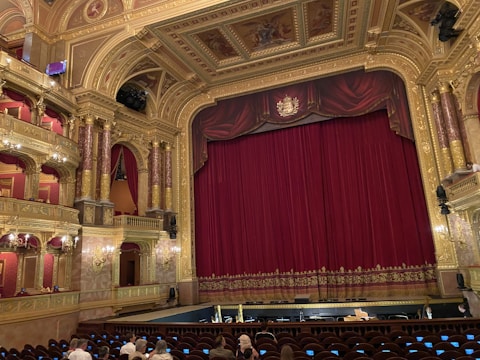 An ornate theatre interior featuring a large red velvet curtain on stage surrounded by gold architectural details. The ceiling and walls are intricately decorated with carvings and paintings, and there are multiple tiers with red velvet seating. A small number of people are seated in the audience area.