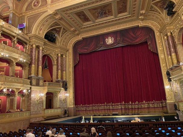 An ornate theatre interior featuring a large red velvet curtain on stage surrounded by gold architectural details. The ceiling and walls are intricately decorated with carvings and paintings, and there are multiple tiers with red velvet seating. A small number of people are seated in the audience area.