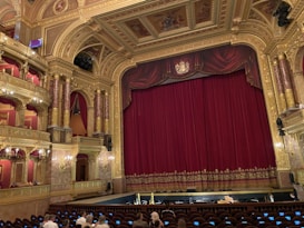 An ornate theatre interior featuring a large red velvet curtain on stage surrounded by gold architectural details. The ceiling and walls are intricately decorated with carvings and paintings, and there are multiple tiers with red velvet seating. A small number of people are seated in the audience area.