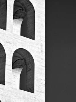 A portion of a modernist building is visible, showcasing a grid of large, round arches set against a stark, dark background. The architecture features smooth, white stone surfaces that create an interplay of light and shadow.