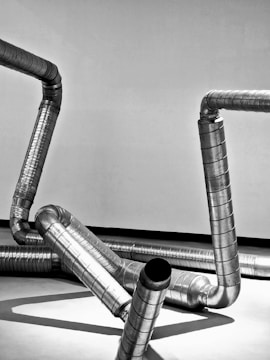 A collection of metallic ventilation ducts are arranged in a twisting and angular pattern against a plain background. The ducts are shiny with reflective surfaces, highlighting the industrial design.
