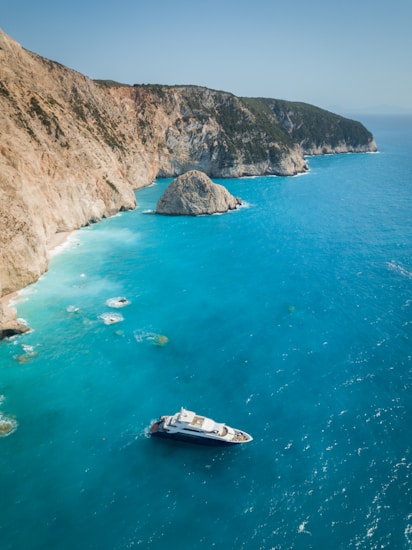A luxurious yacht sails through crystal clear turquoise waters near towering coastal cliffs under a clear blue sky. The rugged cliffs are covered with patches of green vegetation, adding contrast to the rocky surface. A large rock formation protrudes from the water near the shore.