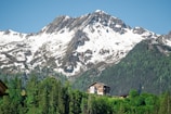 A picturesque scene featuring a snow-capped mountain range with rugged peaks. In the foreground, a cozy wooden chalet surrounded by lush green trees is nestled at the base of the mountains. The clear blue sky above complements the tranquil setting.