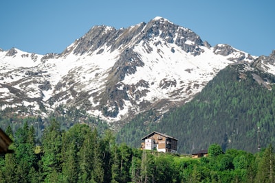 A picturesque scene featuring a snow-capped mountain range with rugged peaks. In the foreground, a cozy wooden chalet surrounded by lush green trees is nestled at the base of the mountains. The clear blue sky above complements the tranquil setting.
