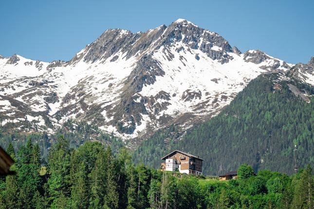 A picturesque scene featuring a snow-capped mountain range with rugged peaks. In the foreground, a cozy wooden chalet surrounded by lush green trees is nestled at the base of the mountains. The clear blue sky above complements the tranquil setting.