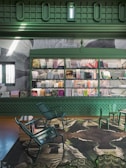 A room with an intricately designed green interior featuring a wall lined with bookshelves filled with various books. The floor hosts chairs and a table primarily made of metal, set on a large, abstract-patterned carpet. The wall above the shelves displays a large artwork of abstract forms.