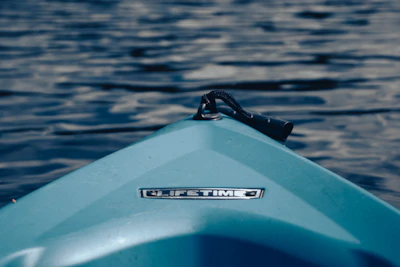 A close-up of the front of a blue kayak on a body of water with gentle ripples. The kayak has a brand label reading 'Lifetime' and a handle attached near the front tip.