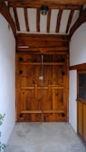 A wooden double door with a rustic appearance sits within a small alcove. The door is made up of vertical wooden planks reinforced with horizontal bars and black metal hinges. Above the door, a ceiling features exposed wooden beams, adding to the traditional aesthetic. The walls surrounding the door are white and stark, contrasting the warm tones of the wood.
