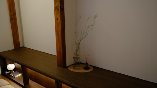 A minimalist interior features a wooden shelf integrated into the wall, with two vertical wooden beams. On the shelf is a glass vase with tall, slender branches and green leaves. Next to the vase is a small incense holder with sticks. Below the shelf, a small lamp and a book are placed, providing a soft glow to the setting.