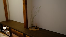 A minimalist interior features a wooden shelf integrated into the wall, with two vertical wooden beams. On the shelf is a glass vase with tall, slender branches and green leaves. Next to the vase is a small incense holder with sticks. Below the shelf, a small lamp and a book are placed, providing a soft glow to the setting.
