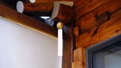 A wooden wall with visible beams and a hanging wind chime. The chime features a brass sphere and a vertical, flat white piece. The setting is in a tranquil corner with natural wood textures accentuated with soft light.