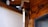 A wooden wall with visible beams and a hanging wind chime. The chime features a brass sphere and a vertical, flat white piece. The setting is in a tranquil corner with natural wood textures accentuated with soft light.